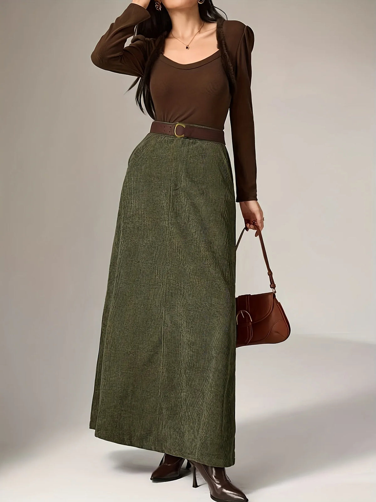 Woman wearing a brown top and green skirt holding a brown handbag on a plain background