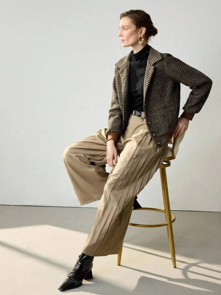 Woman sitting on a stool wearing a plaid jacket, black shirt, beige pants, and black boots in a minimalistic setting.