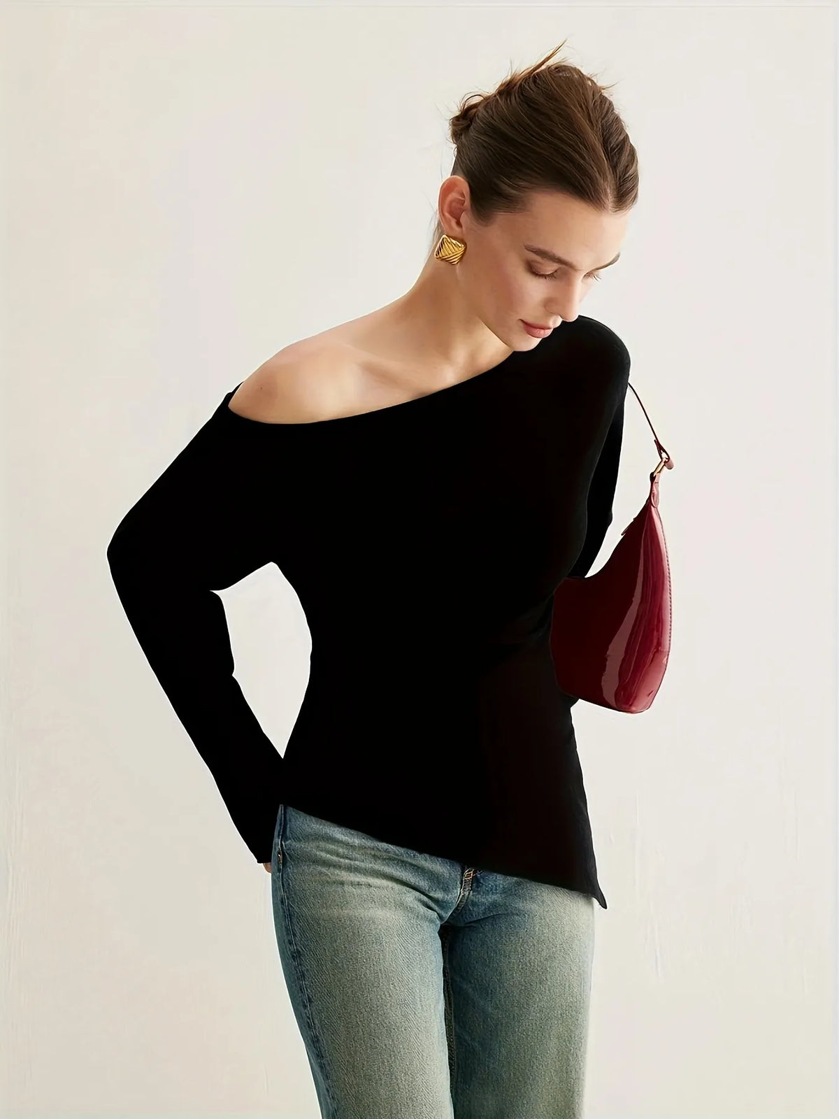 Woman wearing a black off-the-shoulder long-sleeve top paired with light blue jeans. The top has a relaxed fit and an asymmetrical neckline, styled with gold statement earrings and a sleek updo for an elegant minimalist look.