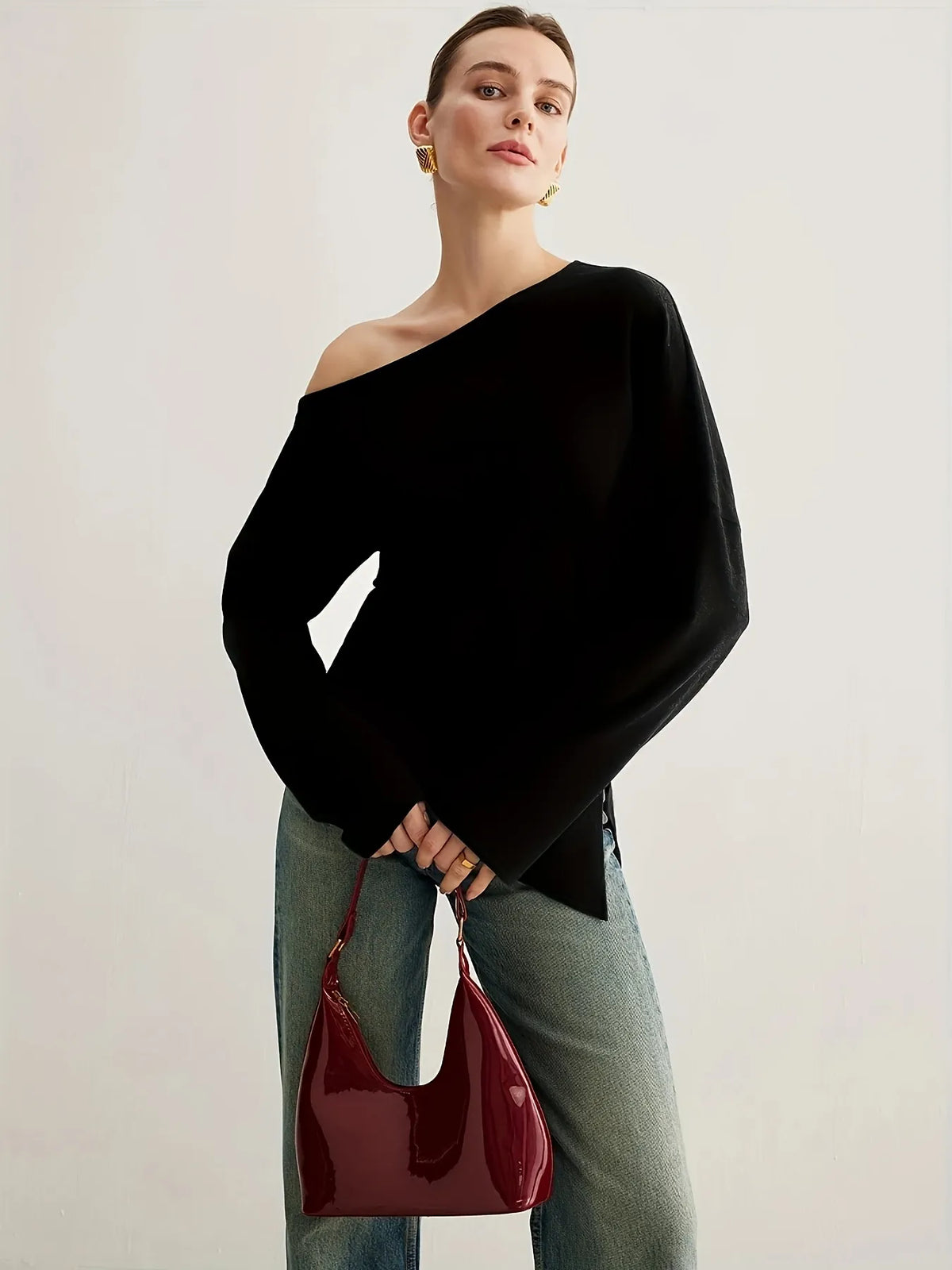Woman wearing a black off-the-shoulder long-sleeve top paired with light blue jeans. The top has a relaxed fit and an asymmetrical neckline, styled with gold statement earrings and a sleek updo for an elegant minimalist look.