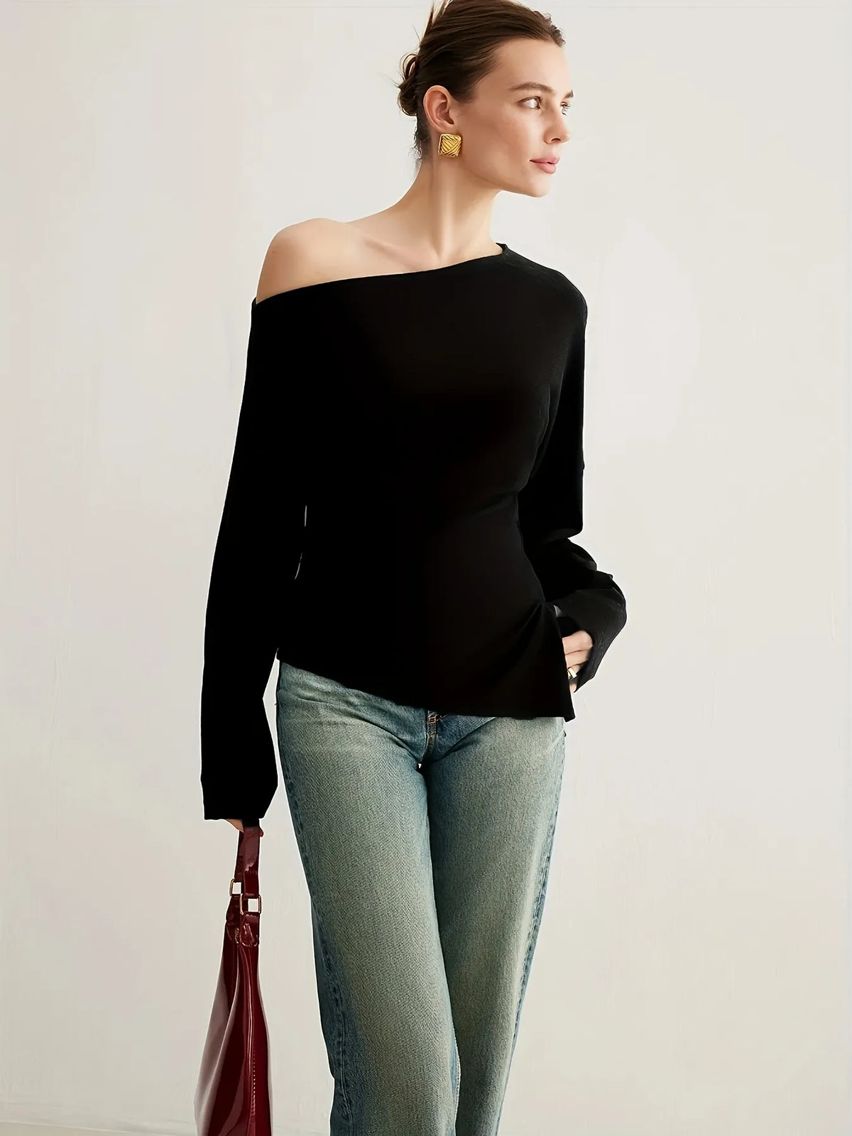 Woman wearing a black off-the-shoulder long-sleeve top paired with light blue jeans. The top has a relaxed fit and an asymmetrical neckline, styled with gold statement earrings and a sleek updo for an elegant minimalist look.