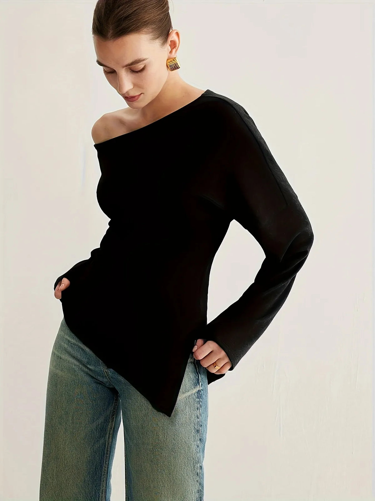 Woman wearing a black off-the-shoulder long-sleeve top paired with light blue jeans. The top has a relaxed fit and an asymmetrical neckline, styled with gold statement earrings and a sleek updo for an elegant minimalist look.