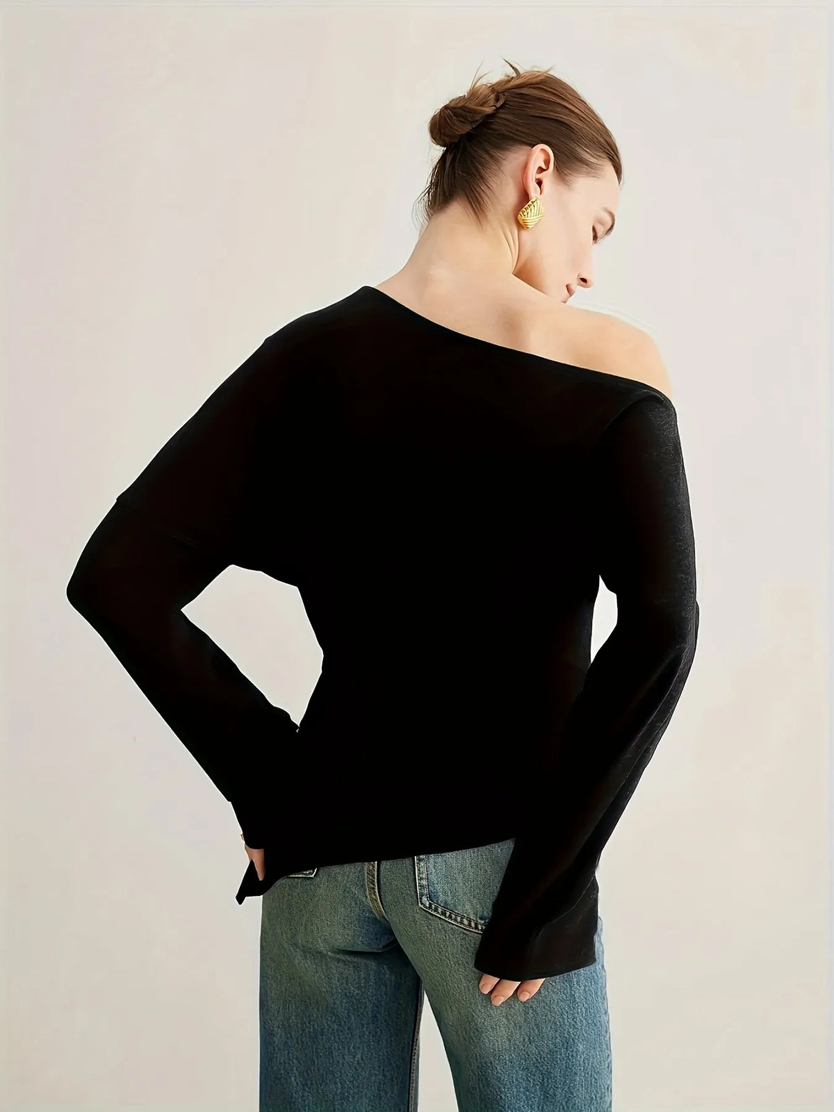 Woman wearing a black off-the-shoulder long-sleeve top paired with light blue jeans. The top has a relaxed fit and an asymmetrical neckline, styled with gold statement earrings and a sleek updo for an elegant minimalist look.