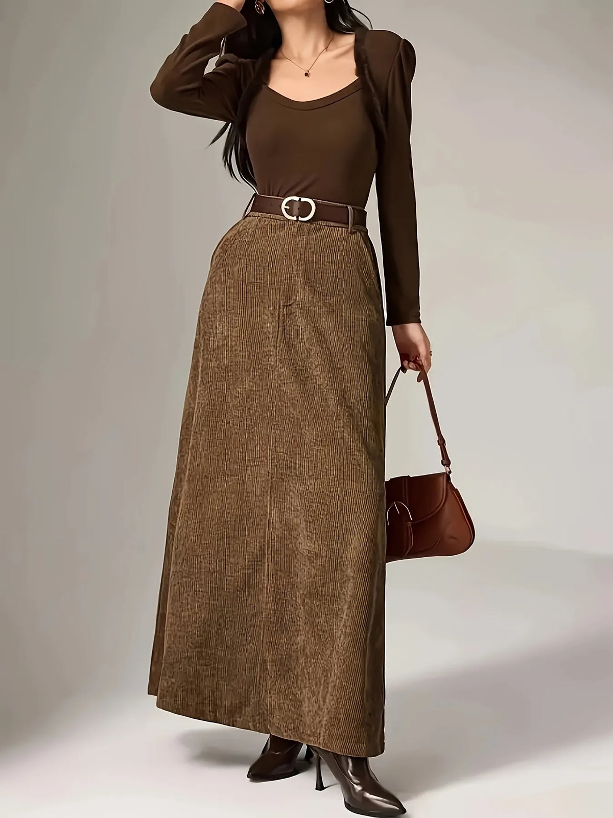 Woman wearing a brown outfit with a long skirt and matching jacket, holding a brown handbag on a plain background.