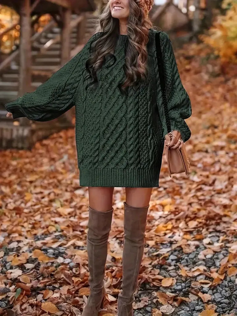 Woman in a green cable knit sweater and brown knee-high boots standing on a leaf-covered ground.