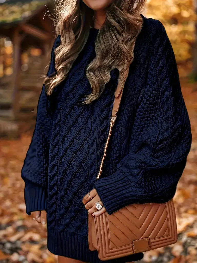 Person wearing a navy blue cable knit sweater with a brown handbag in an autumn setting.
