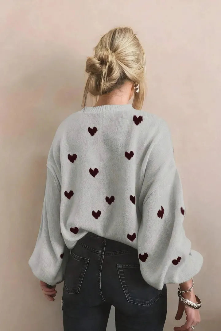Person wearing a light gray sweater with dark red heart patterns on a plain background
