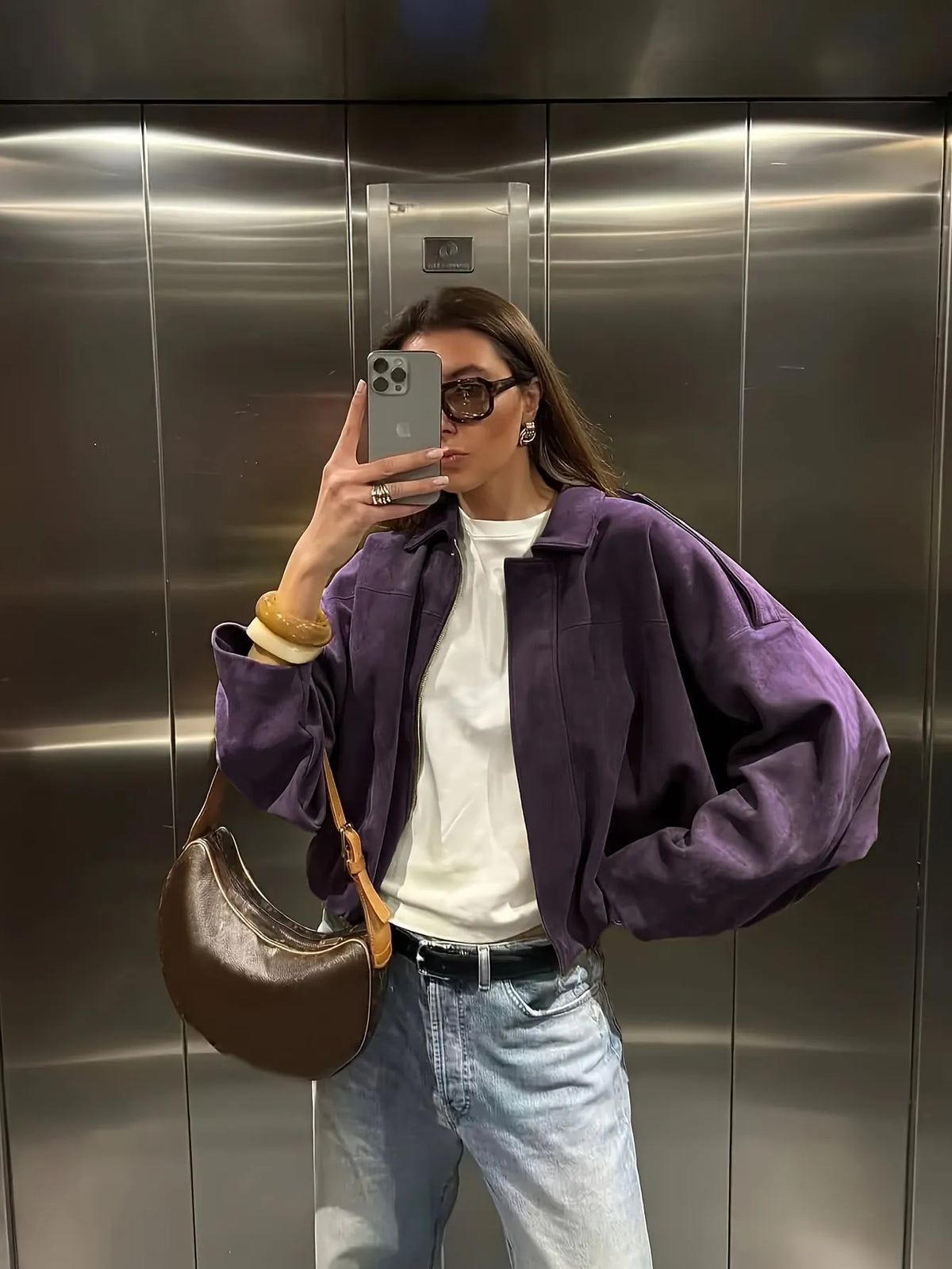 Person taking a mirror selfie wearing a purple jacket, white shirt, blue jeans, and brown bag in an elevator.