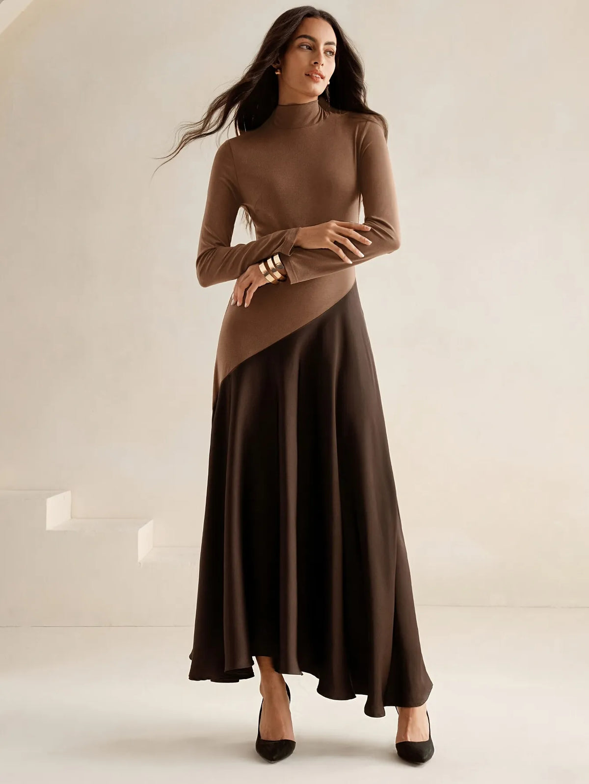 Model wearing a long-sleeve brown high-neck top dress with a diagonal two-tone design and flowing dark brown maxi skirt, paired with black pointed heels, styled in an elegant minimalist look.