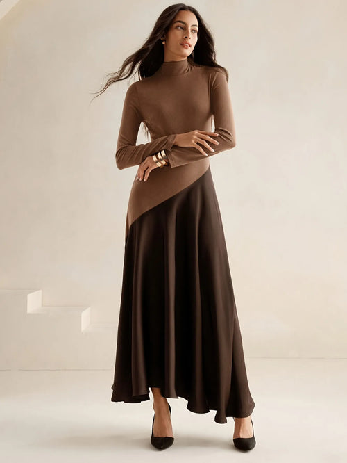 Model wearing a long-sleeve brown high-neck top dress with a diagonal two-tone design and flowing dark brown maxi skirt, paired with black pointed heels, styled in an elegant minimalist look.
