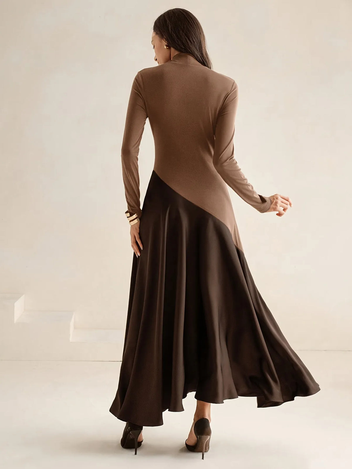 Model wearing a long-sleeve brown high-neck top dress with a diagonal two-tone design and flowing dark brown maxi skirt, paired with black pointed heels, styled in an elegant minimalist look.
