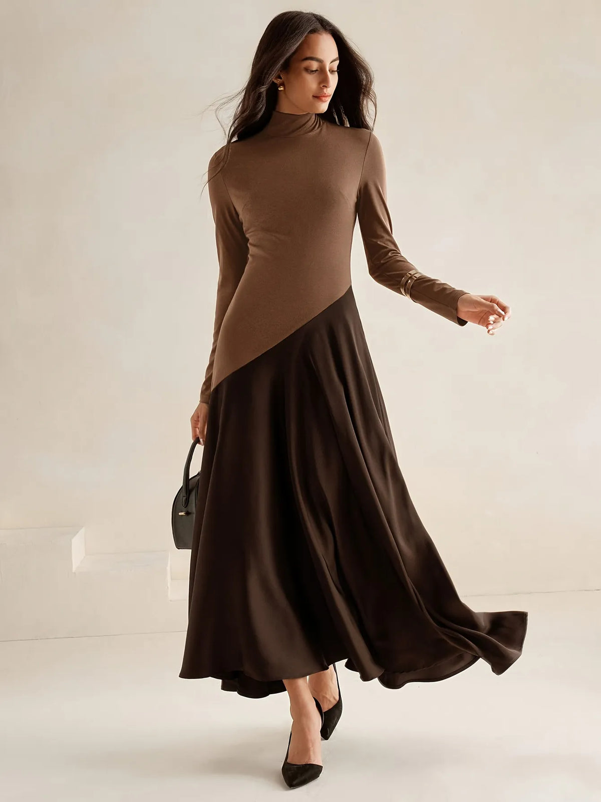 Model wearing a long-sleeve brown high-neck top dress with a diagonal two-tone design and flowing dark brown maxi skirt, paired with black pointed heels, styled in an elegant minimalist look. 