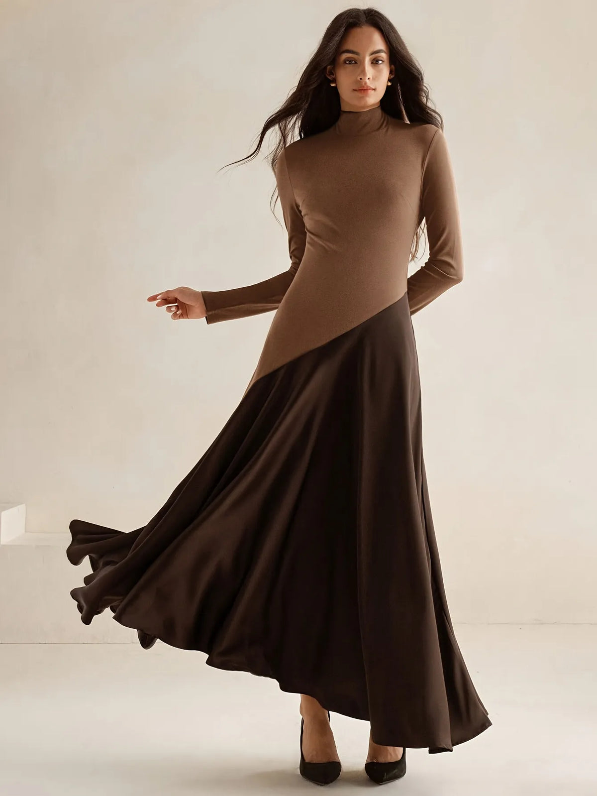 Model wearing a long-sleeve brown high-neck top dress with a diagonal two-tone design and flowing dark brown maxi skirt, paired with black pointed heels, styled in an elegant minimalist look. 