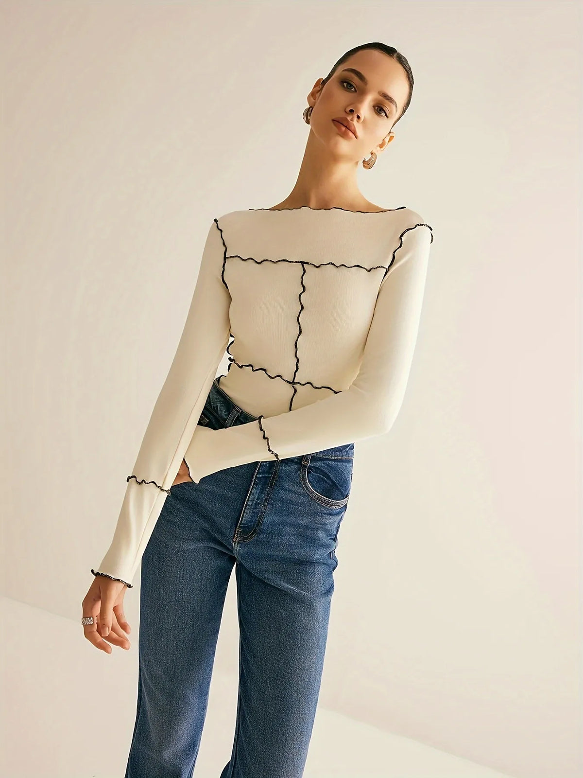 Woman wearing a cream long-sleeve top with black contrast wavy stitching and a fitted silhouette, paired with high-waisted blue jeans. The look is completed with hoop earrings and a sleek bun hairstyle for a modern minimalist aesthetic.