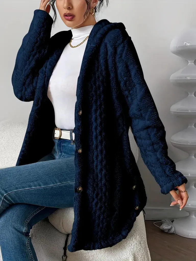 Person wearing a navy blue textured coat over a white top and blue jeans, sitting on a white stool.