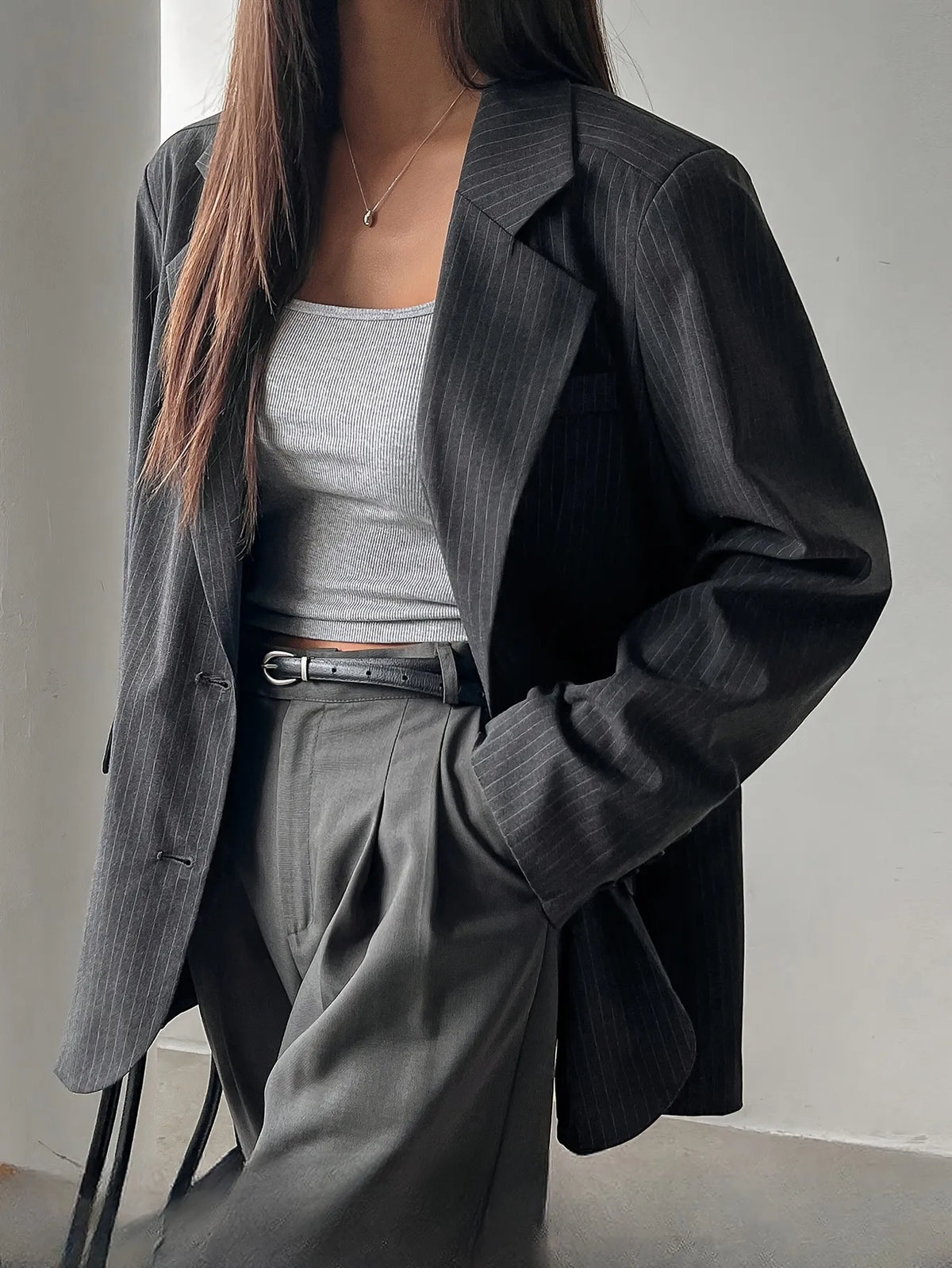 Person wearing a black blazer over a gray top and pants in a minimalistic setting