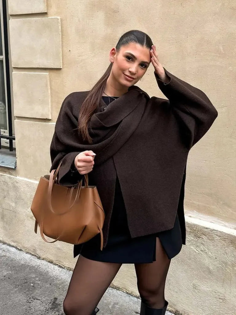 Woman wearing a brown poncho and holding a brown leather bag against a beige wall.