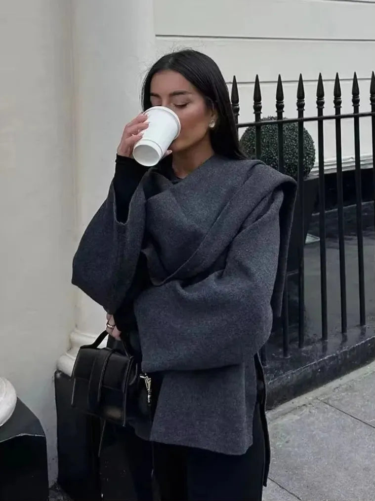 Woman drinking from a white cup while wearing a dark gray coat in an urban setting.