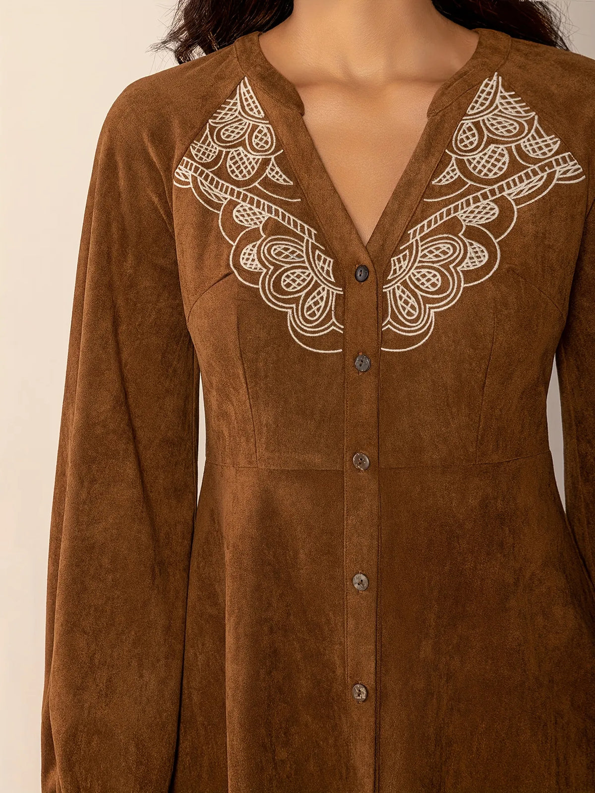 
Woman wearing a short brown button-up dress with long puffed sleeves and embroidered lace detail on the chest. The dress has a relaxed fit and a soft suede-like texture, styled with simple gold hoop earrings.