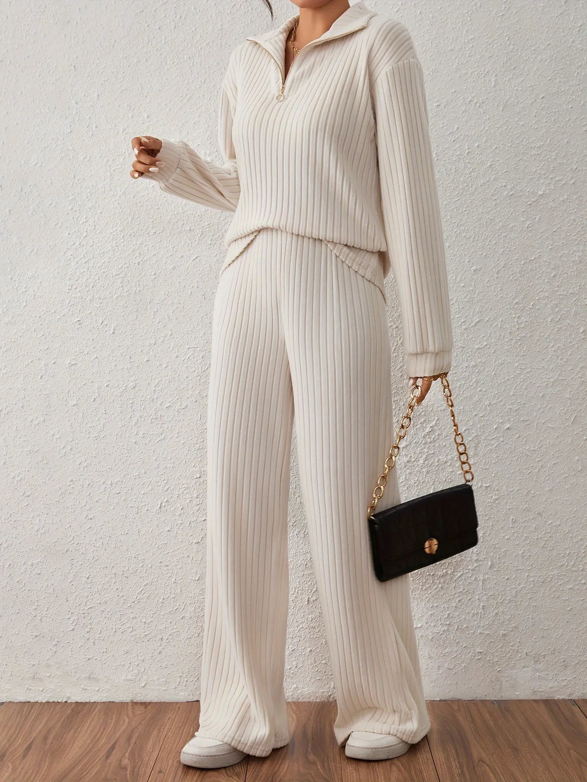 Person wearing a beige ribbed knit outfit holding a black handbag against a plain background