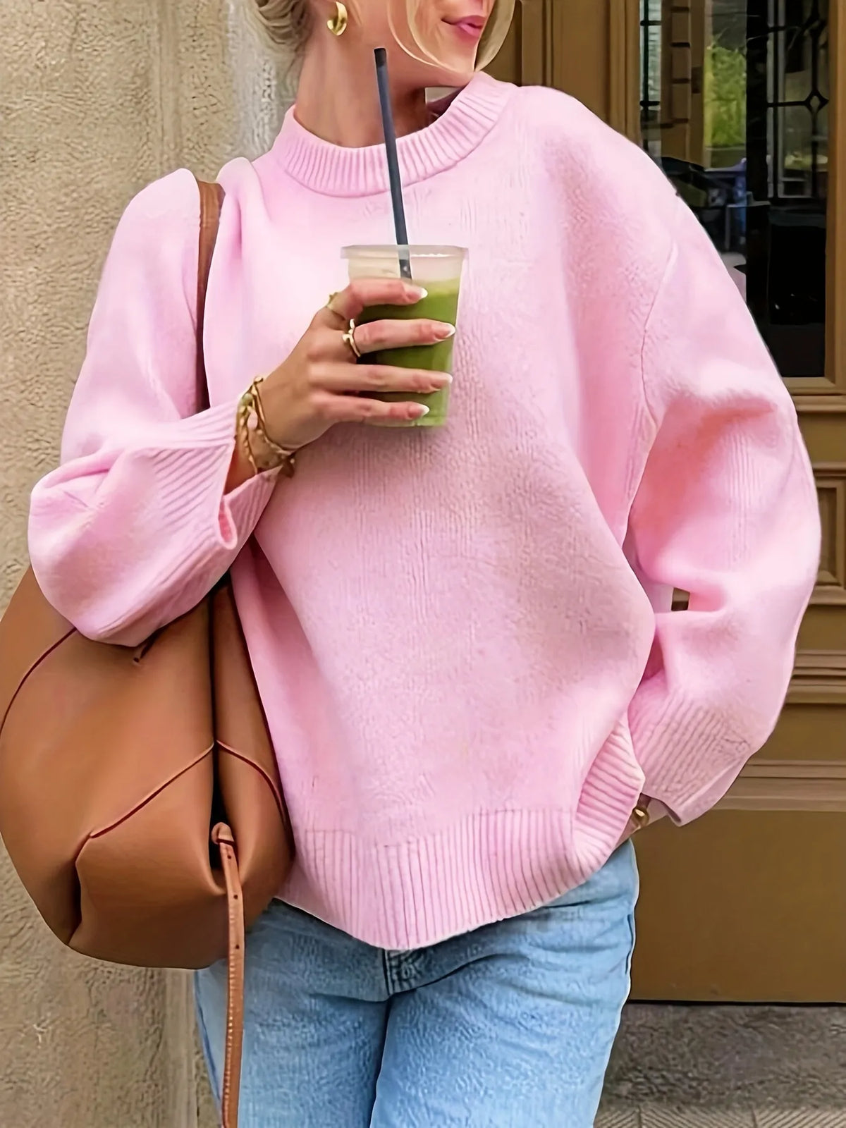 Person wearing a pink sweater holding a green drink, standing against a neutral wall.
