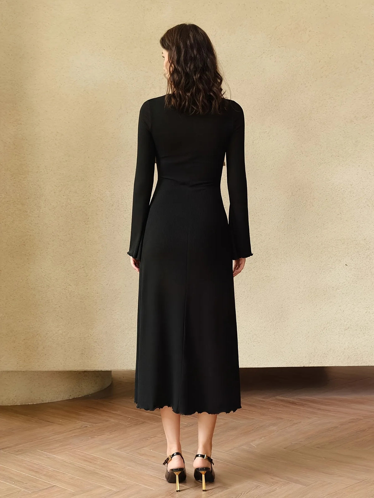 Woman wearing a long black dress with beige twist-front detail and flared sleeves. The dress has a fitted silhouette with a contrasting bodice design, styled with subtle gold hoop earrings and loose wavy hair.