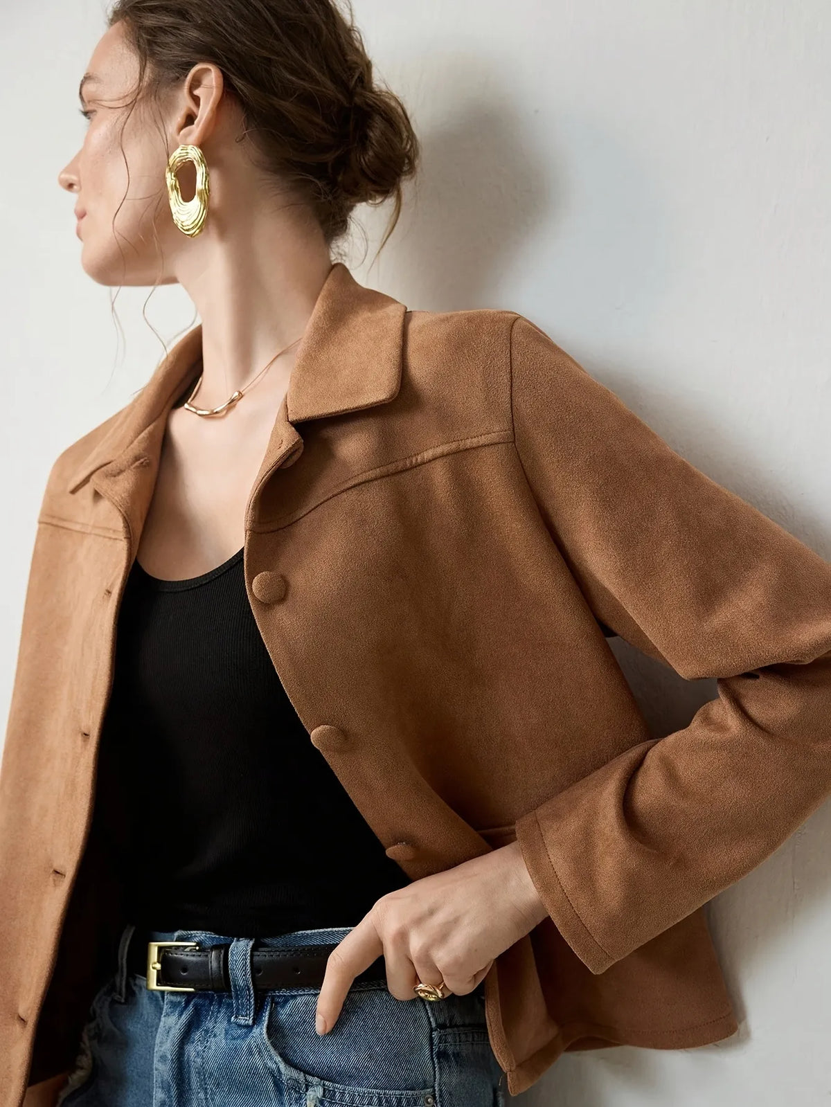 Woman wearing a cropped camel-brown suede jacket with button details, paired with a black ribbed tank top, blue jeans, and a black belt. She accessorises with large gold hoop earrings and a gold necklace, her hair styled in a loose bun.