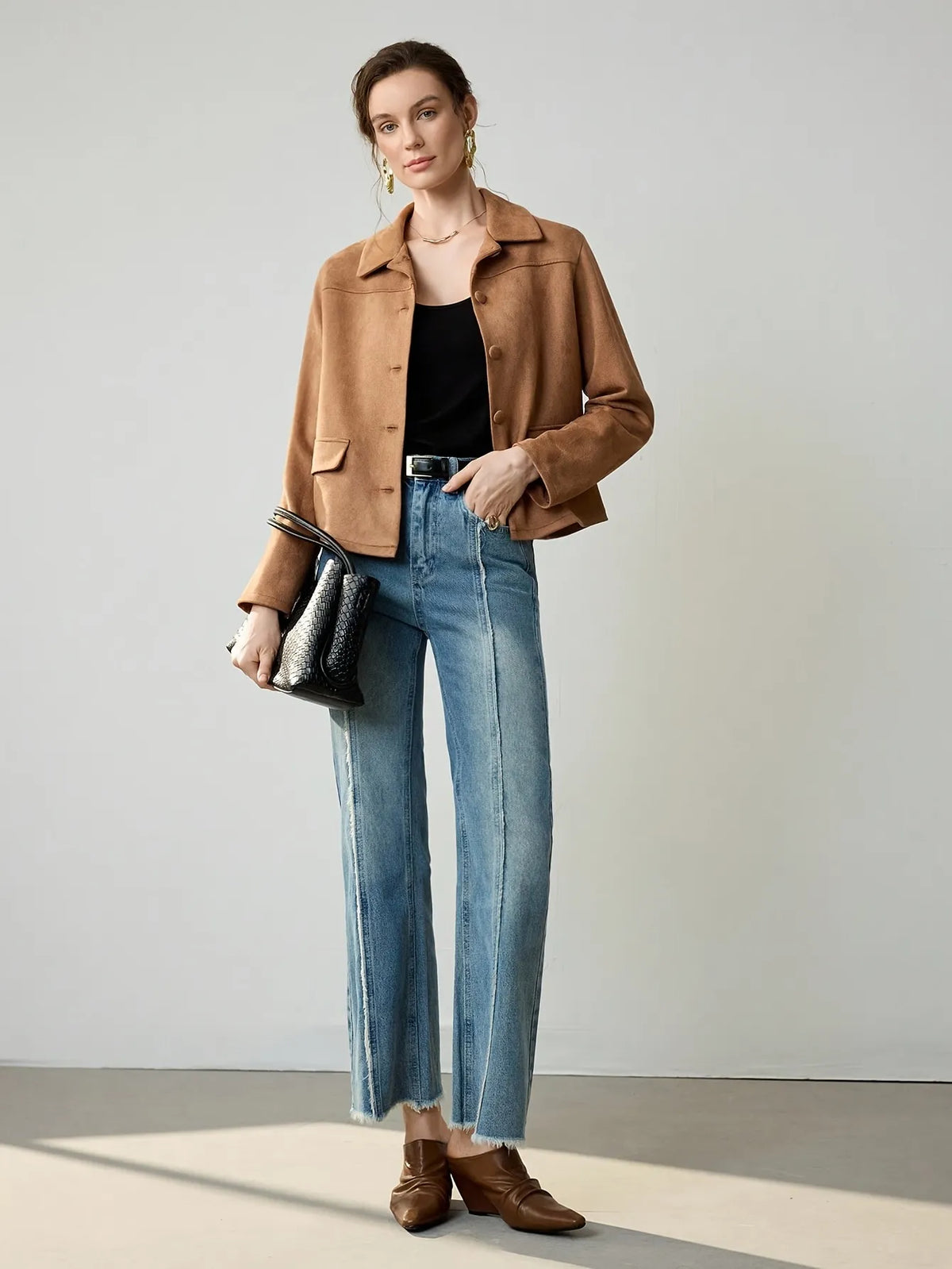 Woman wearing a cropped camel-brown suede jacket with button details, paired with a black ribbed tank top, blue jeans, and a black belt. She accessorises with large gold hoop earrings and a gold necklace, her hair styled in a loose bun.