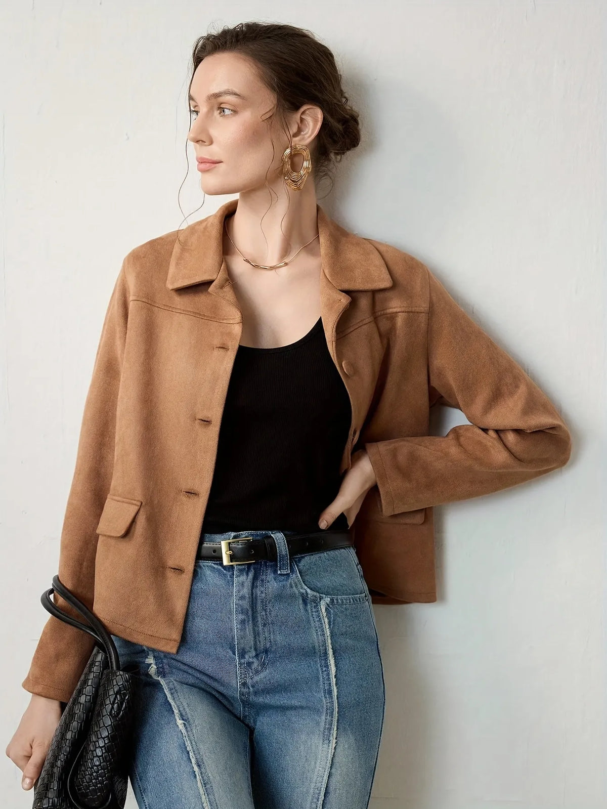 Woman wearing a cropped camel-brown suede jacket with button details, paired with a black ribbed tank top, blue jeans, and a black belt. She accessorises with large gold hoop earrings and a gold necklace, her hair styled in a loose bun.