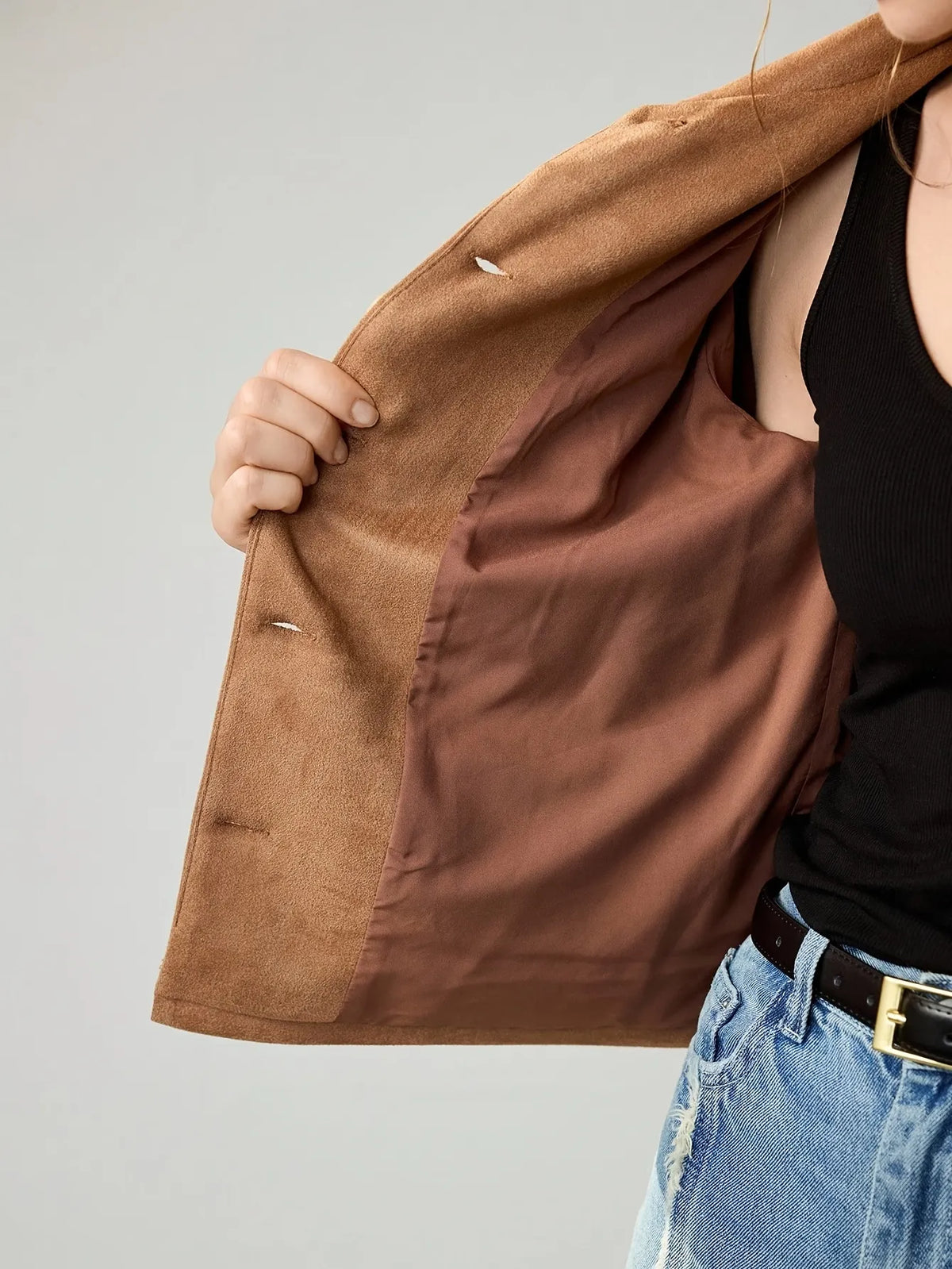 Woman wearing a cropped camel-brown suede jacket with button details, paired with a black ribbed tank top, blue jeans, and a black belt. She accessorises with large gold hoop earrings and a gold necklace, her hair styled in a loose bun.